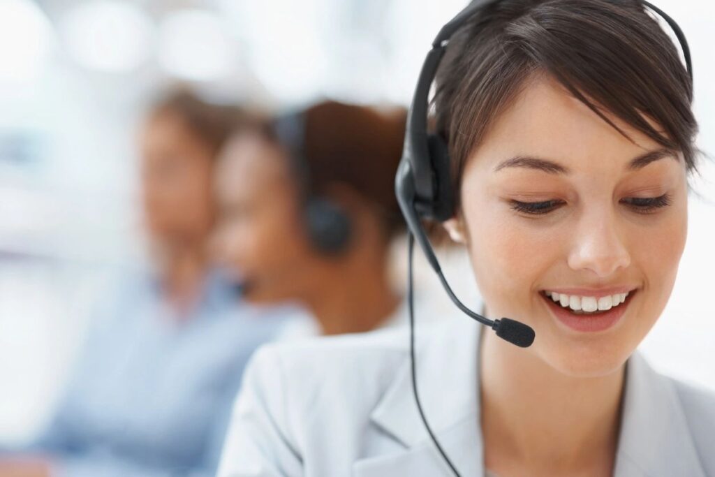 Contact Centers Phone System: Young woman with a headset, smiling at her desk in a contact center, with two blurred colleagues assisting customers in the background.