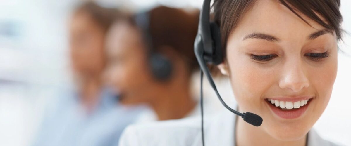 Contact Centers Phone System: Young woman with a headset, smiling at her desk in a contact center, with two blurred colleagues assisting customers in the background.