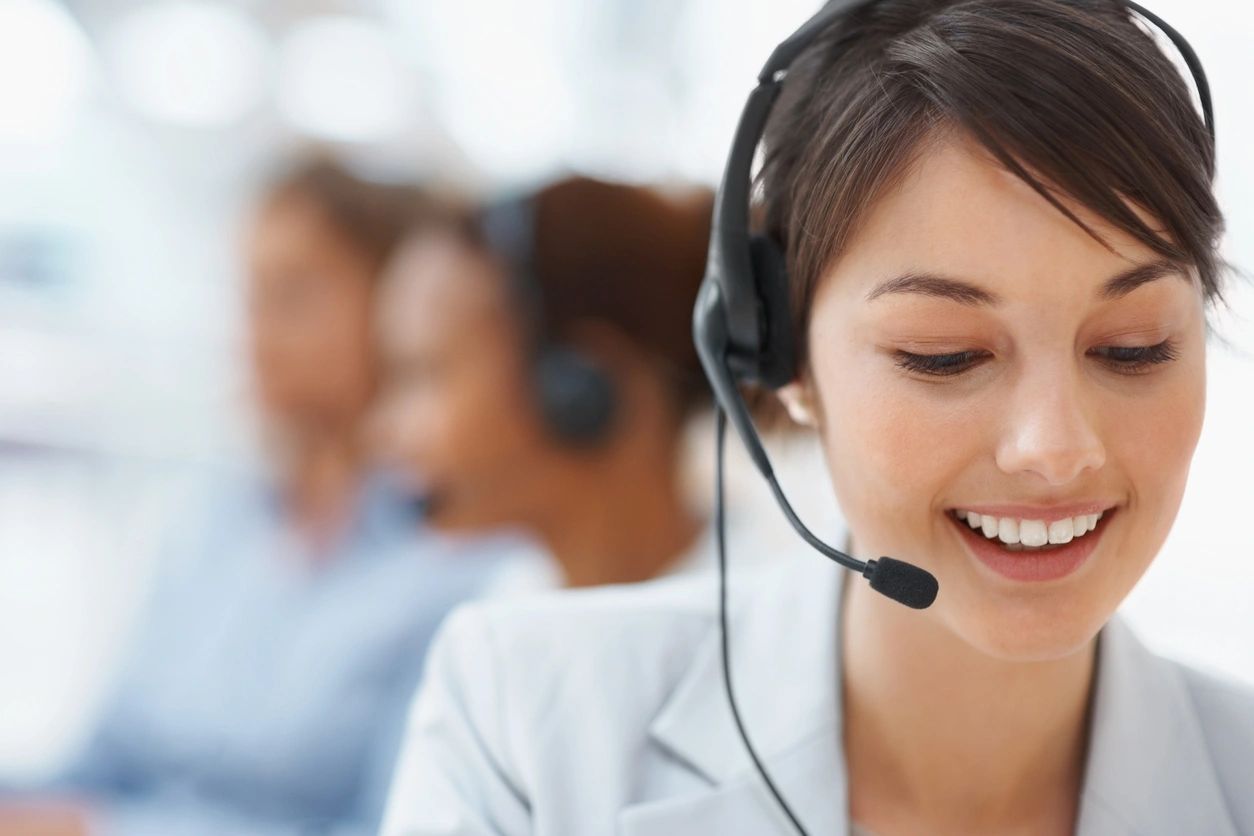 Contact Centers Phone System: Young woman with a headset, smiling at her desk in a contact center, with two blurred colleagues assisting customers in the background.